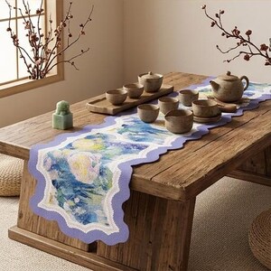 Pode incluir: Um caminho de mesa retangular com uma borda recortada em lavanda e uma estampa central com padrões florais abstratos em azul, verde e amarelo. O caminho está sobre uma mesa de madeira ao lado de um bule de cerâmica e pequenas xícaras.