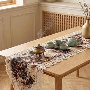 Op de afbeelding: Een rechthoekige tafelloper met een donker bloemenpatroon en crèmekleurige kanten randen. De loper ligt op een houten tafel met een gouden wierookbrander, drie kleine lichtgroene theekopjes, een bijpassende theepot en een gouden vaas.