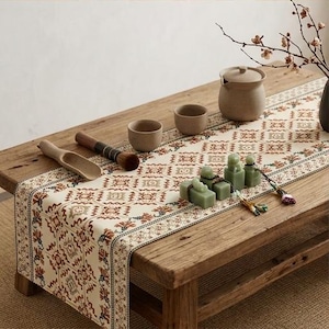 Puede incluir: Un camino de mesa color crema con un patrón geométrico y floral repetitivo en tonos óxido, marrón y verde oscuro. El camino de mesa se exhibe sobre una mesa de madera junto a dos cuencos de cerámica pequeños, una tetera y sellos de piedra verde.