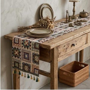 Puede incluir: Un camino de mesa rectangular con un patrón geométrico repetitivo de cuadrados en tonos marrón, crema, azul y rojo. El camino de mesa está rematado con pequeñas borlas colgantes en colores verde y beige alternados en el borde inferior.