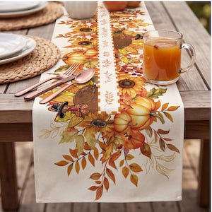 May include: A cream-colored fabric table runner featuring a vibrant autumn-themed print of sunflowers, orange pumpkins, and turkeys wearing pilgrim hats, accented by a central vertical strip with a checkered border and leaf patterns.