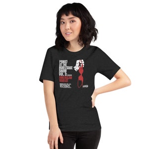 May include: Dark gray t-shirt featuring the text "PANIC! AT THE BURLESQUE SHOW VOL. 3: PRESENTS AMERICAN HARLOT" in white and red. The design includes a stylized illustration of a hand holding a bra. The shirt also has event details.