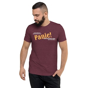 Può includere: T-shirt bordeaux con la scritta "Panic!" in un carattere arancione stilizzato. La maglietta include anche il testo "at the burlesque show" e i dettagli dell'evento. La maglietta è indossata da una persona.