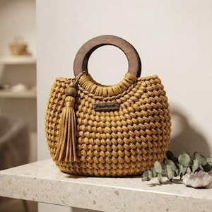 Puede incluir: Un bolso de ganchillo color amarillo mostaza con un asa redonda de madera oscura, una borla a juego sujeta en el lateral y una pequeña etiqueta rectangular con la palabra Handmade impresa en la parte frontal del tejido texturizado.