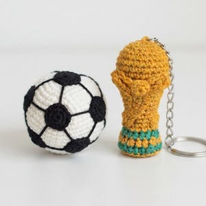 May include: A crocheted soccer ball with a white and black geometric pattern alongside a gold-colored crocheted trophy keychain with a green base and a silver metal ring, both crafted with visible yarn stitches and a textured finish.