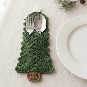 May include: A dark green crocheted Christmas tree shaped cutlery holder with a brown base and a small white star at the top. The holder contains a silver metal fork and a silver metal spoon, positioned vertically within the crocheted pocket.