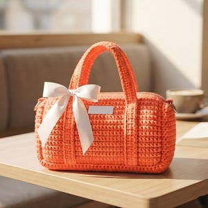 May include: A bright orange crochet handbag featuring a rectangular structure with two top handles and a white satin ribbon bow tied to the side. The bag has a small, blank white rectangular label centered on the front panel and metal hardware.