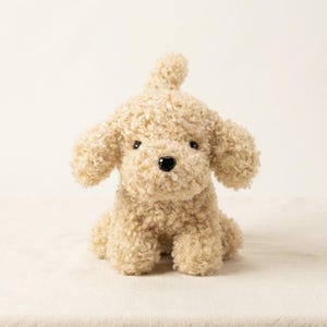 Op de afbeelding: Een knuffel in de vorm van een kleine hond met krullende, lichtbeige vacht. Het knuffeldier heeft een ronde kop, hangende oren, een kleine zwarte neus en donkere ogen. Het is afgebeeld in een zittende houding met de staart omhoog.