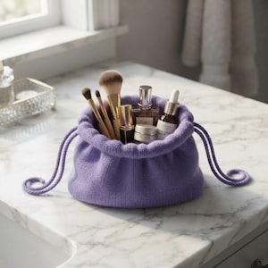 May include: A lavender knit drawstring pouch containing several makeup brushes, a small glass perfume bottle, a cylindrical cosmetic container, and a dropper bottle. The pouch features a thick, textured knit fabric and matching purple cord ties.