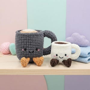 Könnte beinhalten: Zwei gehäkelte Tassen-Plüschtiere mit lächelnden Gesichtern. Die größere Tasse ist dunkelgrau mit beigen Füßen und Schaumkrone. Die kleinere Tasse ist weiß mit dunkelbraunen Füßen und Schaumkrone. Beide haben schwarze Augen und rosa Wangen.
