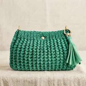 May include: A rectangular emerald green crochet handbag featuring a thick cord texture, a central gold-tone metal snap button closure, and a matching green leather-like tassel attached to a gold-tone metal ring on the right side of the bag.