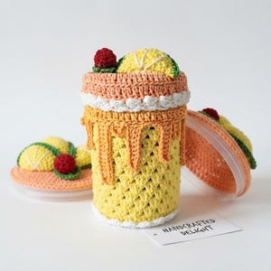 May include: A crochet-covered cylindrical container featuring a yellow textured base, a peach-colored top with white scalloped trim, and a lid decorated with a yellow lemon slice and a red berry. A white tag reads HANDCRAFTED DELIGHT.