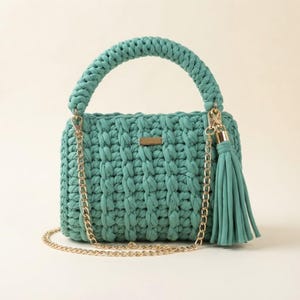 Peut inclure: Un sac à main en crochet bleu sarcelle doté d'une anse tressée épaisse, d'une bandoulière en chaîne métallique dorée et d'un pompon assorti. L'avant du sac présente une petite plaque métallique rectangulaire dorée centrée en haut.
