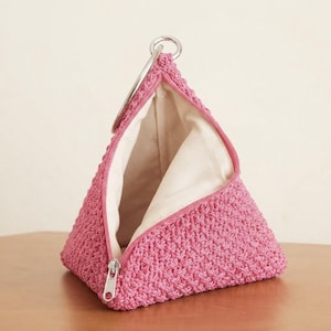 May include: A triangular pouch crafted from pink crocheted yarn featuring a silver-toned metal zipper closure and a circular metal ring handle at the top. The interior is lined with a plain off-white fabric, and the structure is rigid.