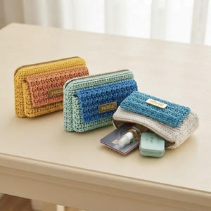 Peut inclure: Trois pochettes rectangulaires en crochet avec des fermetures éclair en métal doré et une petite étiquette métallique rectangulaire marquée PARDA. Les pochettes sont jaunes, bleues et blanches, chacune avec une poche à rabat. Une pochette est ouverte, révélant une carte bancaire, un petit vaporisateur et un récipient rectangulaire vert clair.