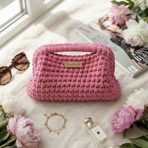 May include: A pink crocheted handbag with a structured frame and a gold-toned metal plaque on the front that reads "home made". The bag features a thick, textured knit pattern and a top handle, presented as a singular accessory item.