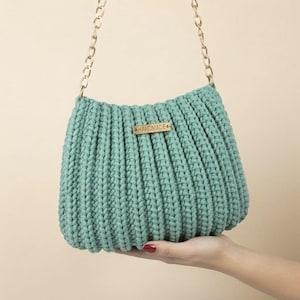 May include: A sage green crocheted handbag featuring a vertical ribbed texture and a gold-toned metal chain strap. A small rectangular wooden tag with the word HANDMADE engraved in black is attached to the center front of the bag.