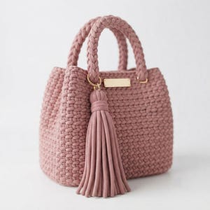 Peut inclure: Un sac à main rose poudré fabriqué en cordon tressé épais avec un motif tissé texturé. Le sac présente deux anses tressées et un grand pompon assorti attaché à un anneau doré. Une plaque métallique rectangulaire dorée est centrée.