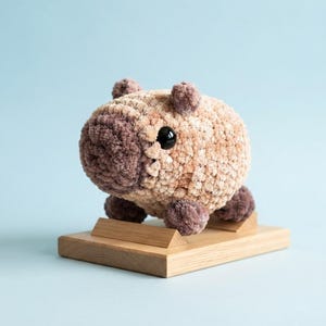 Pode incluir: Uma pelúcia de capivara feita em crochê com corpo de fio texturizado em tons de bege e marrom, pequenas orelhas arredondadas e um olho de plástico preto visível. A pelúcia está montada sobre uma base retangular de madeira de cor clara.