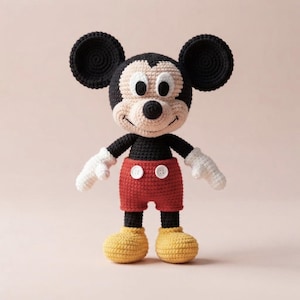 May include: A crochet doll of a cartoon mouse character with large round black ears, a black head, and a cream-colored face. The figure wears red shorts with two white buttons, white gloves, and yellow shoes, all crafted with a textured yarn stitch.