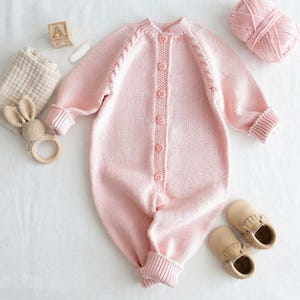 Può includere: Una tutina in maglia a maniche lunghe color rosa chiaro con un'abbottonatura centrale dotata di sei bottoni coordinati, polsini a coste e un dettaglio a treccia lungo le maniche raglan, presentata con un gomitolo di lana e scarpine.