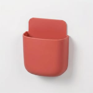May include: A terracotta-coloured wall-mounted storage pocket with a smooth, matte finish. The container features a rounded rectangular shape with an open top and a flat, extended back panel designed for attachment to a vertical surface.
