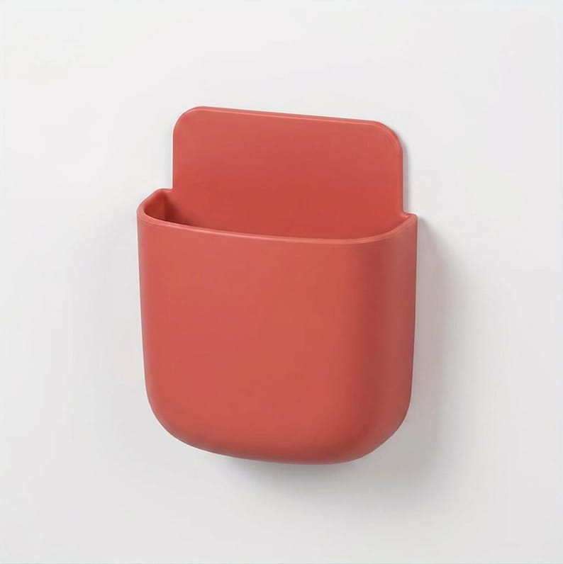 May include: A terracotta-coloured wall-mounted storage pocket with a smooth, matte finish. The container features a rounded rectangular shape with an open top and a flat, extended back panel designed for attachment to a vertical surface.