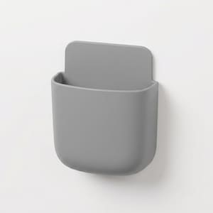 May include: A matte grey wall-mounted storage pocket with a rounded rectangular shape and a flat back panel. The container features a smooth, solid-coloured surface with an open top, designed for holding small items in a vertical orientation.