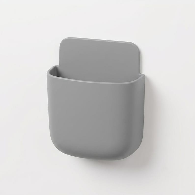 May include: A matte grey wall-mounted storage pocket with a rounded rectangular shape and a flat back panel. The container features a smooth, solid-coloured surface with an open top, designed for holding small items in a vertical orientation.