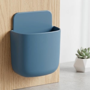 May include: A matte blue wall-mounted storage pocket with a rounded rectangular shape and a flat back panel. The container features a smooth, solid-coloured surface and an open top, designed to be attached to a vertical surface for holding small items.