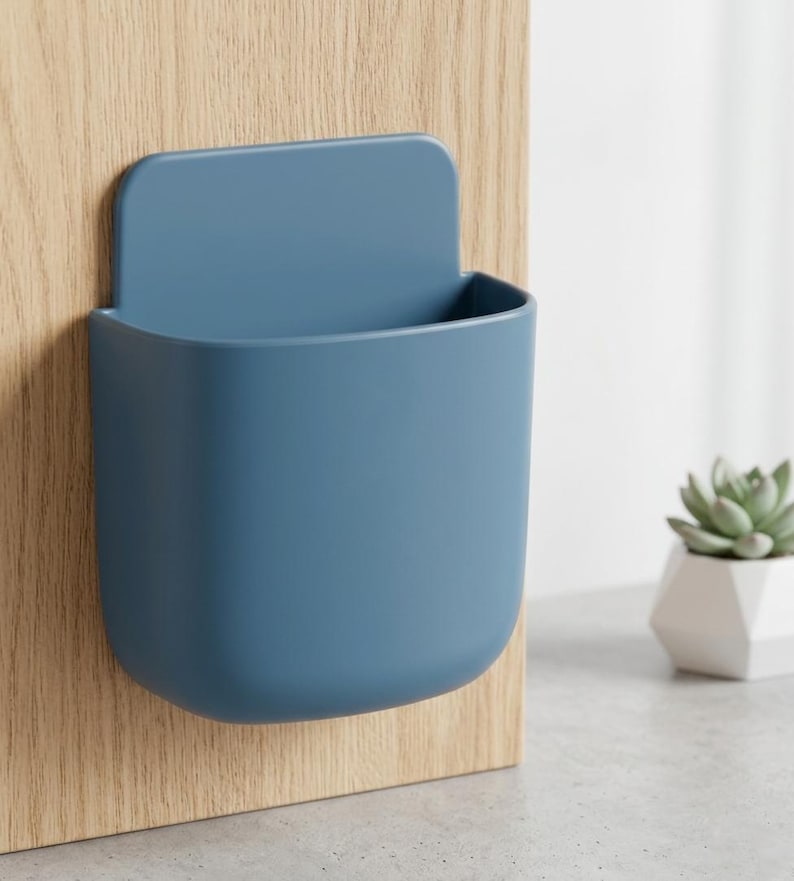 May include: A matte blue wall-mounted storage pocket with a rounded rectangular shape and a flat back panel. The container features a smooth, solid-coloured surface and an open top, designed to be attached to a vertical surface for holding small items.