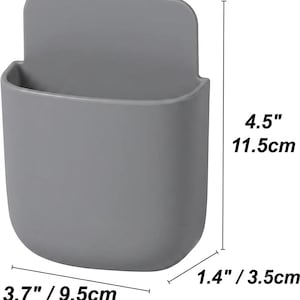 May include: A grey wall-mounted storage pocket with a rounded rectangular shape. The dimensions are clearly marked as 11.5 cm in height, 9.5 cm in width, and 3.5 cm in depth. The container features a smooth, solid-coloured surface.