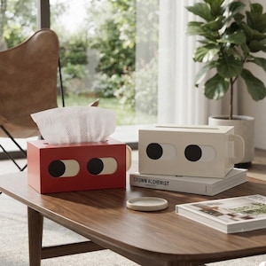 May include: Two rectangular tissue box covers, one red and one off-white, each featuring a stylised face design with two horizontal oval eyes. The red cover holds a white tissue, while the off-white cover sits atop a book labelled Grown Alchemist.