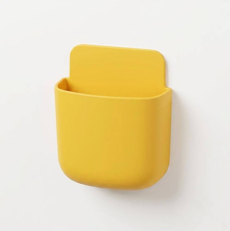 May include: A bright yellow wall-mounted storage pocket with a smooth, matte finish. This U-shaped container features a flat back panel for attachment and a deep, open-top compartment designed for holding small items securely against a flat surface.