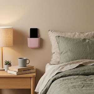 May include: A matte light pink wall-mounted storage pocket holding a black smartphone. The rectangular container features rounded corners and a smooth finish, designed to be attached to a flat vertical surface for holding small electronic devices.