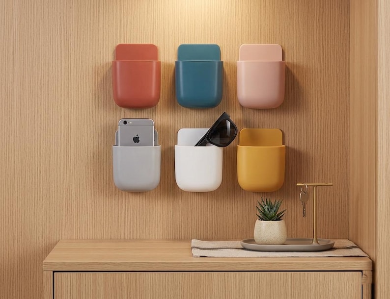 May include: Six wall-mounted storage pockets in terracotta, teal, light pink, grey, white, and mustard yellow. The grey pocket holds a silver smartphone, and the white pocket contains black sunglasses. Each pocket has a flat back and rounded base.