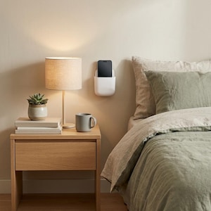 May include: A white wall-mounted phone holder with a rectangular pocket design, securely attached to a neutral-coloured wall above a wooden nightstand. The holder contains a black smartphone, positioned vertically within the storage compartment.