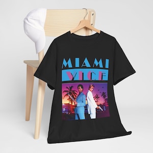 Puede incluir: Una camiseta negra de manga corta con un estampado gráfico que incluye el texto MIAMI VICE en azul y rosa. El gráfico muestra a dos figuras con traje frente a un atardecer con palmeras. También se ve una gorra de béisbol blanca.
