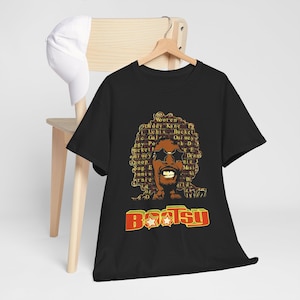 May include: A black short-sleeve t-shirt featuring a graphic of a person with an afro hairstyle composed of various names, wearing star-shaped sunglasses. Below the graphic, the word BOOTSY is printed in large red and yellow block letters.
