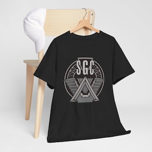 May include: A black short-sleeve t-shirt featuring a grey circular graphic on the chest with the letters SGC centered above a stylized triangular emblem with wing-like details. A plain white baseball cap is draped over the back of the chair.