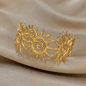 Peut inclure: Un bracelet manchette doré présentant un motif ajouré répétitif de soleils stylisés avec des centres en spirale et des rayons pointus rayonnants. Le métal a une finition polie et le bracelet est formé en cercle ouvert pour permettre l'ajustement.