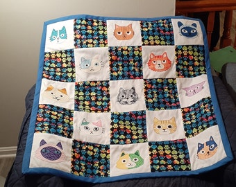 May include: A blue and white baby quilt with a patchwork design featuring various cat faces embroidered on each square. The quilt has a blue border and a black and white background with hearts and paw prints.