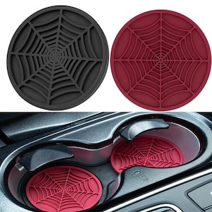 May include: Two circular cup holder inserts featuring a raised spider web pattern. One insert is black and the other is deep red. The red inserts are shown placed inside a vehicle's dual cup holder console, providing a protective base for drinks.