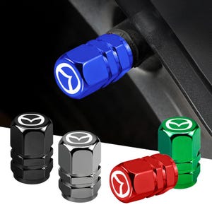 May include: A collection of cylindrical tyre valve stem caps in blue, black, silver, red, and green. Each cap features a hexagonal grip pattern and a white Mazda logo printed on the top surface. One blue cap is shown installed on a tyre valve.