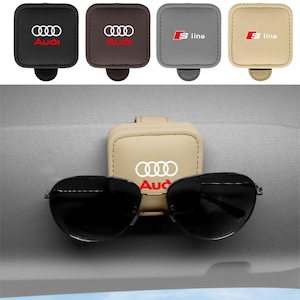 May include: A square leather car visor sunglasses holder featuring the Audi logo in white and red. The holder is available in black, brown, grey, and beige. One beige holder is shown attached to a car visor, securely holding black sunglasses.