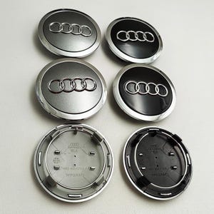 May include: Six circular Audi wheel centre caps arranged in two columns. Four caps show the front with silver or black finishes and the silver Audi four-ring logo. Two caps show the black plastic underside with moulded text including Germany and 4B0 601 170A.