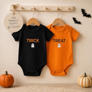 May include: Two short-sleeve baby bodysuits hanging on wooden hangers. One is black with the orange text TRICK and a white ghost icon. The other is orange with the black text TREAT and a white ghost icon. A round sign reads THE PERFECT OUTFIT for Twins!