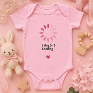 May include: A light pink baby bodysuit featuring a dark grey loading icon graphic, the text "Baby Girl Loading.." and "SEE YOU IN 2027", and a small dark grey heart icon centred on the chest. The garment has short sleeves and three snaps.