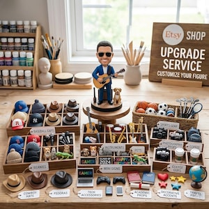 May include: A custom bobblehead figure of a person in a blue suit playing an acoustic guitar, accompanied by a small dog. The scene includes various miniature accessories organized in wooden trays, labeled with tags like baseball caps, instruments, and pets.