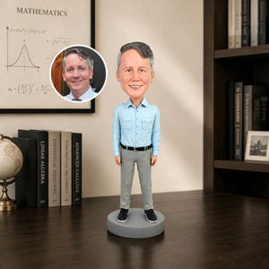 Op de afbeelding: Een gepersonaliseerde bobblehead-figuur van een persoon met grijs haar, gekleed in een lichtblauw overhemd met twee borstzakken, een zwarte riem, een grijze broek en zwarte schoenen, staand op een ronde grijze voet met een ronde foto-inzet.